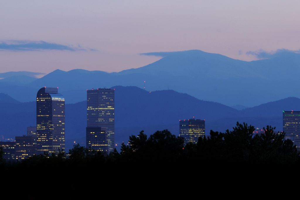 The Denver skyline at dusk with the Rocky Mountains in the background, buildings illuminated against the evening sky.