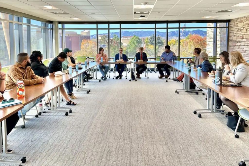 People seated around a large conference table, engaged in a discussion, with a view of the mountains through large windows in the background.