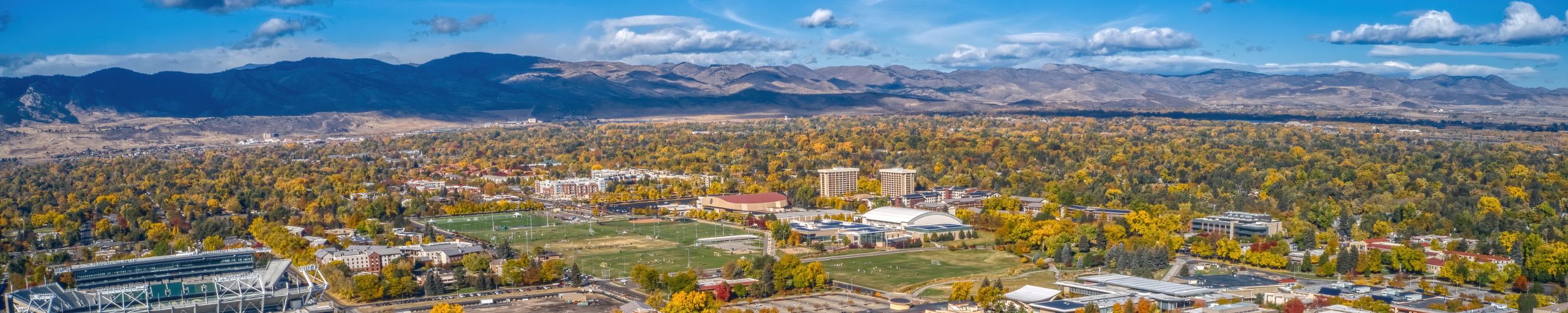 Aerial view of the Colorado State University campus, surrounded by autumn trees, with the Rocky Mountains in the background under a bright blue sky.