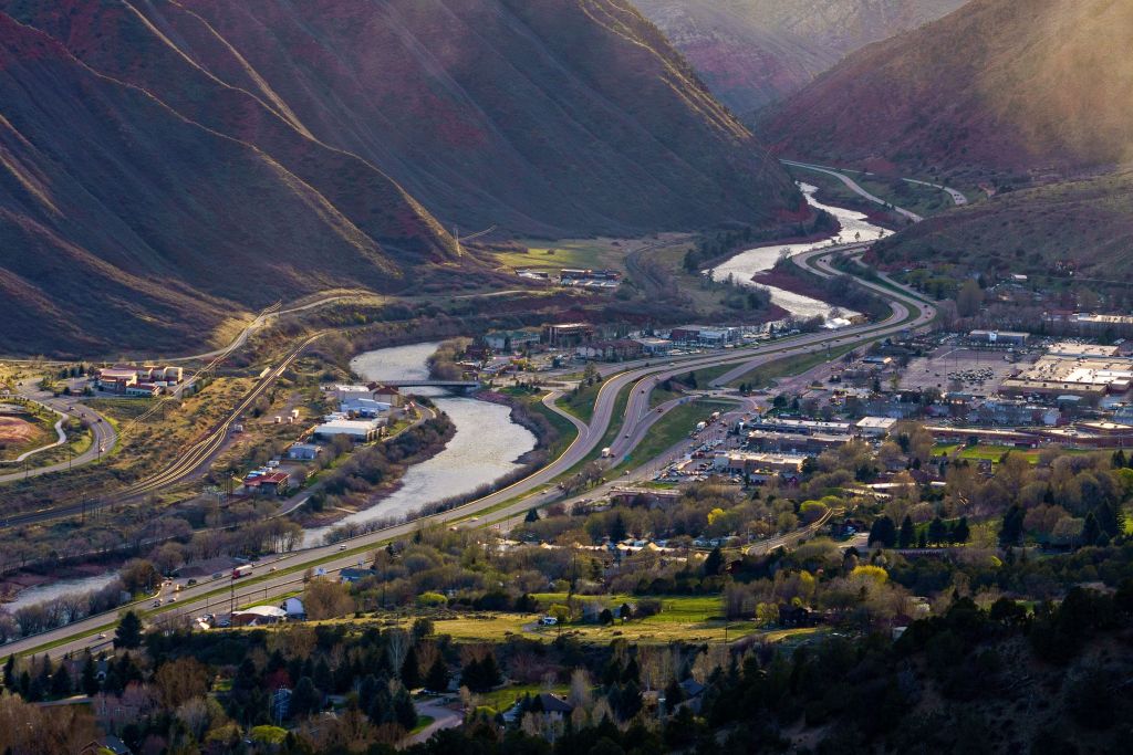 An aerial view of a town nestled in a valley, surrounded by winding roads, rivers, and lush green mountains under warm sunlight.