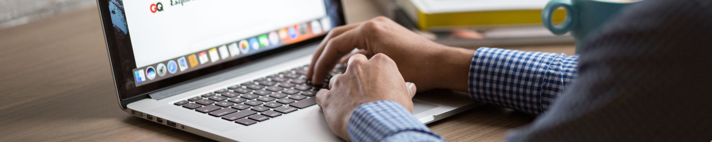 Close-up of a person's hands typing on a laptop keyboard, with a colorful coffee mug and books in the background.