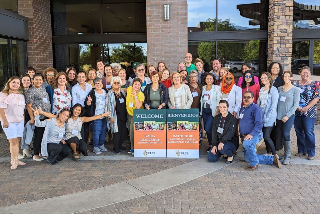 A large group of diverse participants from the Family Leadership Training Institute posing for a group photo outside a building, holding bilingual welcome banners in English and Spanish.