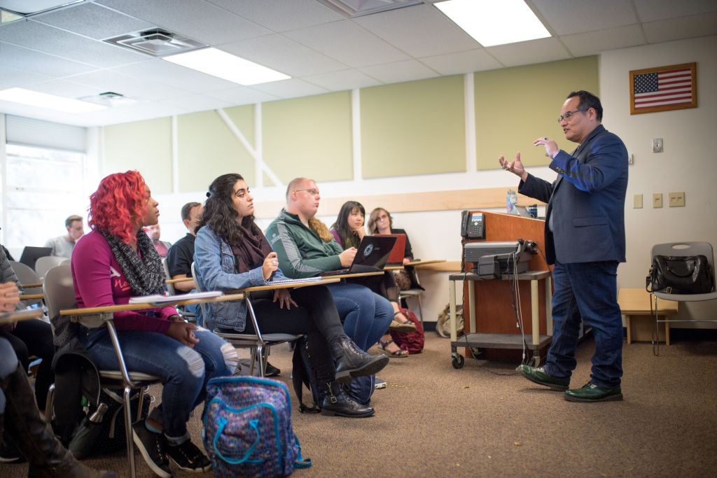 A diverse classroom of students attentively listening to a male professor who is giving a lecture, with desks, chairs, and an American flag in the room.