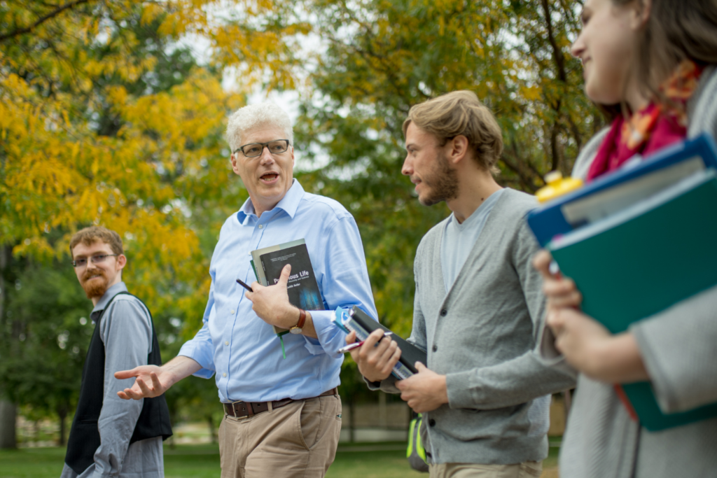 A professor walking and talking with a group of students on a college campus during autumn, with fall foliage in the background.