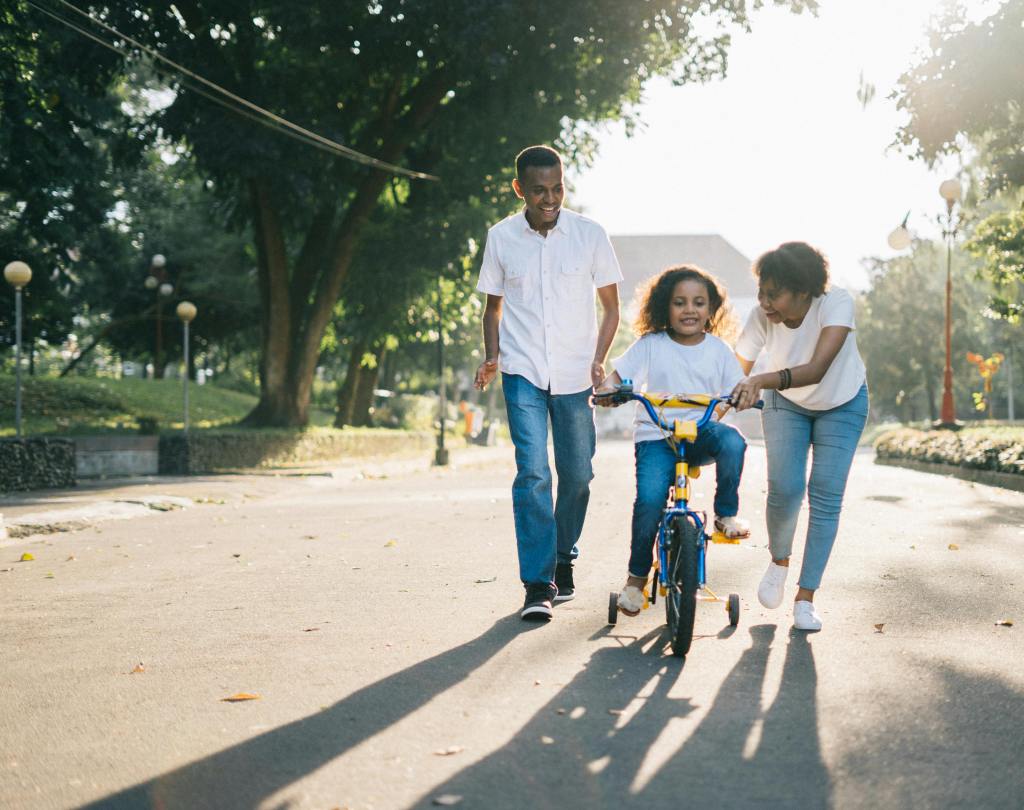 A child learning to ride a bike with the help of two adults on a sunny day.