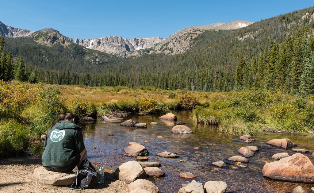 Person wearing a CSU sweatshirt sitting on a rock by a river, with mountains in the background.