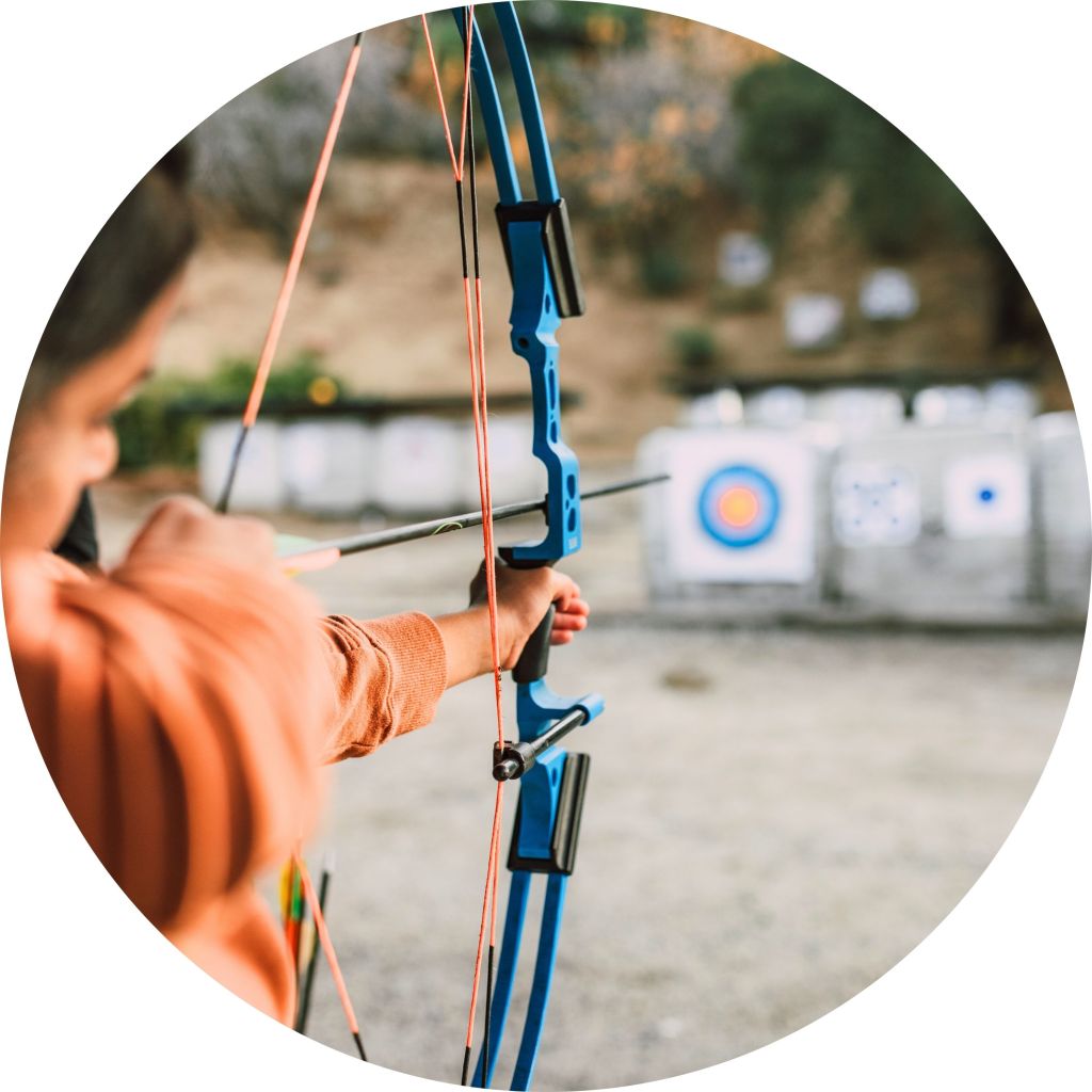 A close-up of a young person holding a blue bow and arrow, aiming at archery targets in the distance. The background shows several targets, including one with a bullseye, suggesting a practice session at an outdoor archery range.