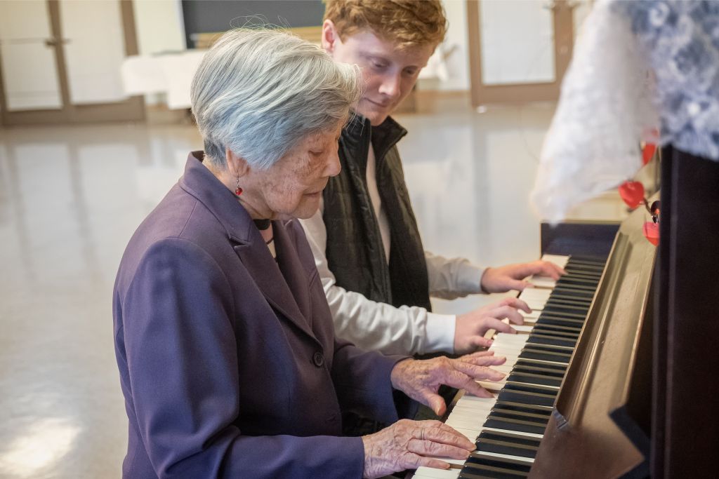 A young man and an elderly woman play the piano together, creating a heartwarming moment of intergenerational music-making.