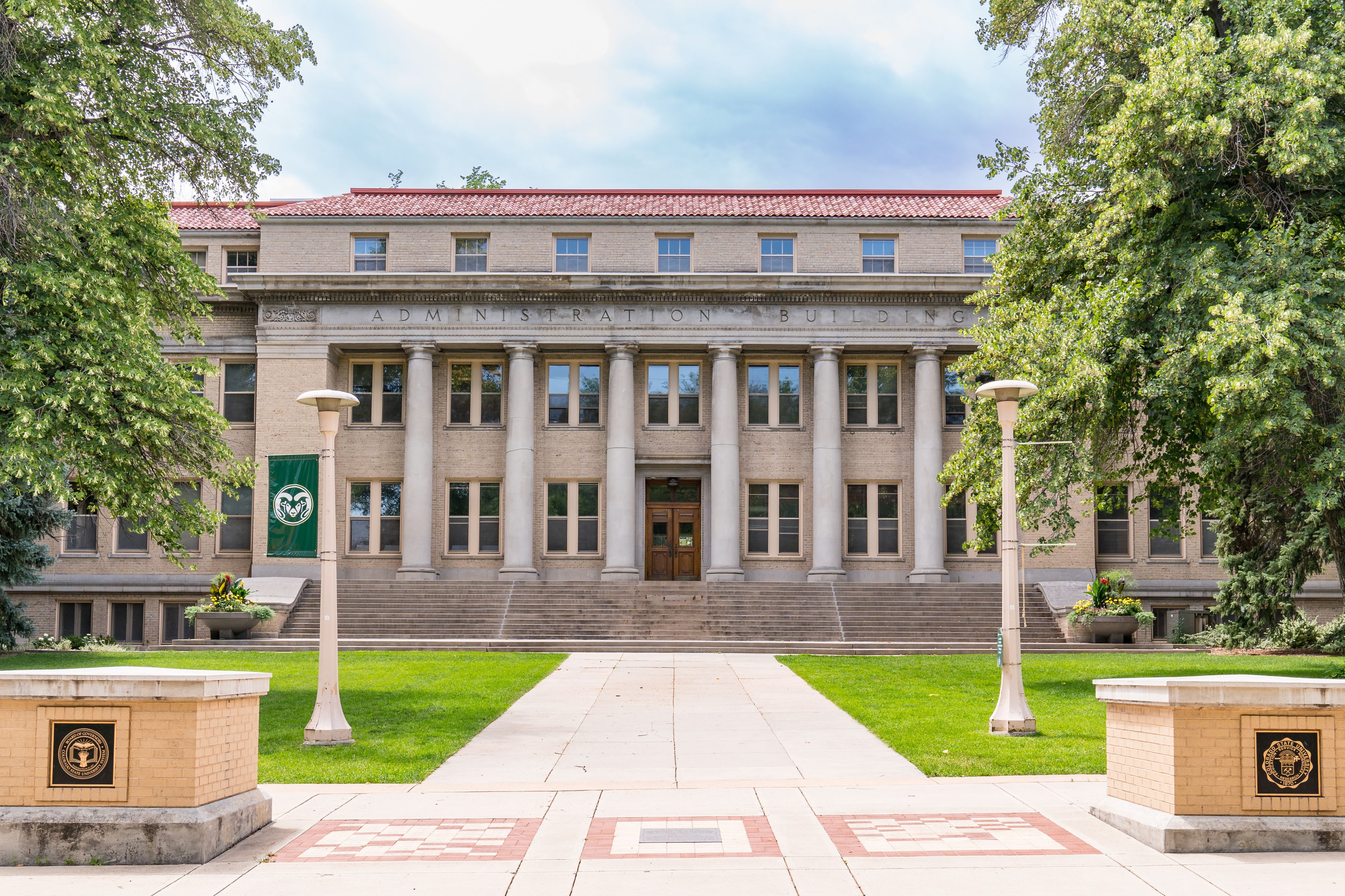 The Colorado State University Administration Building with tall columns, a red-tiled roof, and green CSU banners, framed by green trees and a pathway leading to the entrance.