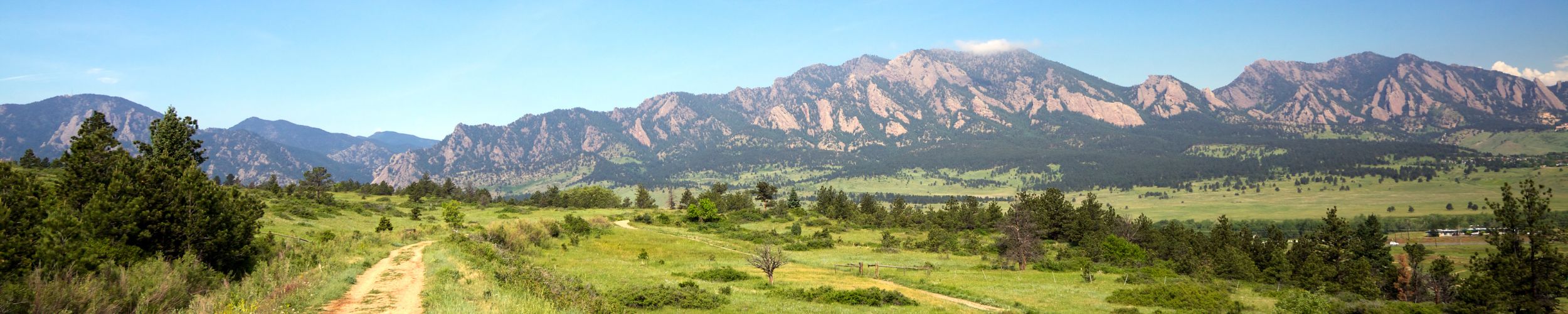 A panoramic view of the Rocky Mountains with grassy fields and a dirt trail in the foreground.