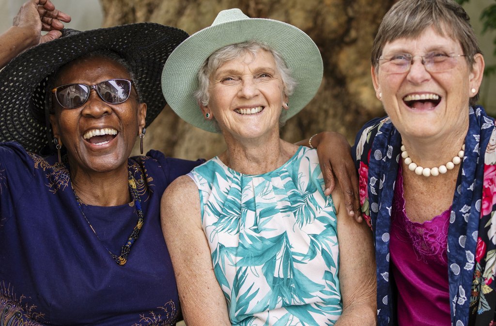 Three elderly women are laughing together, wearing hats and enjoying a sunny day, showcasing friendship and joy in an outdoor setting.