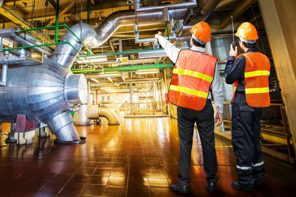 Two engineers wearing safety vests and hard hats are inspecting large industrial machinery inside a factory setting.