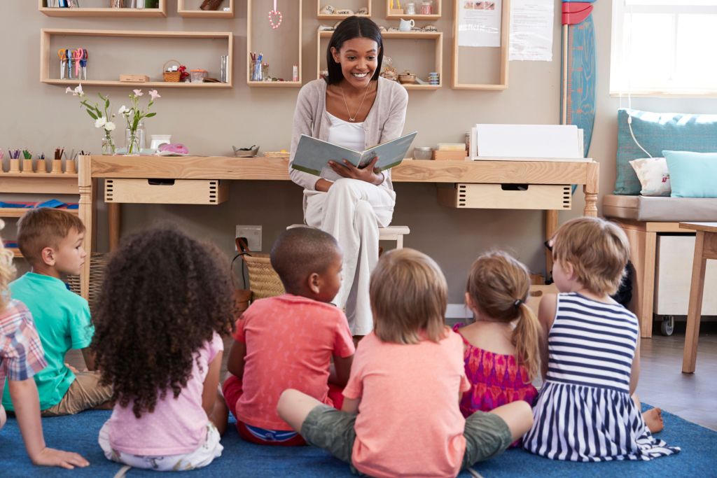 A smiling teacher reading a book to a group of young children sitting on the floor in a classroom, showcasing an engaging learning environment for early childhood education.