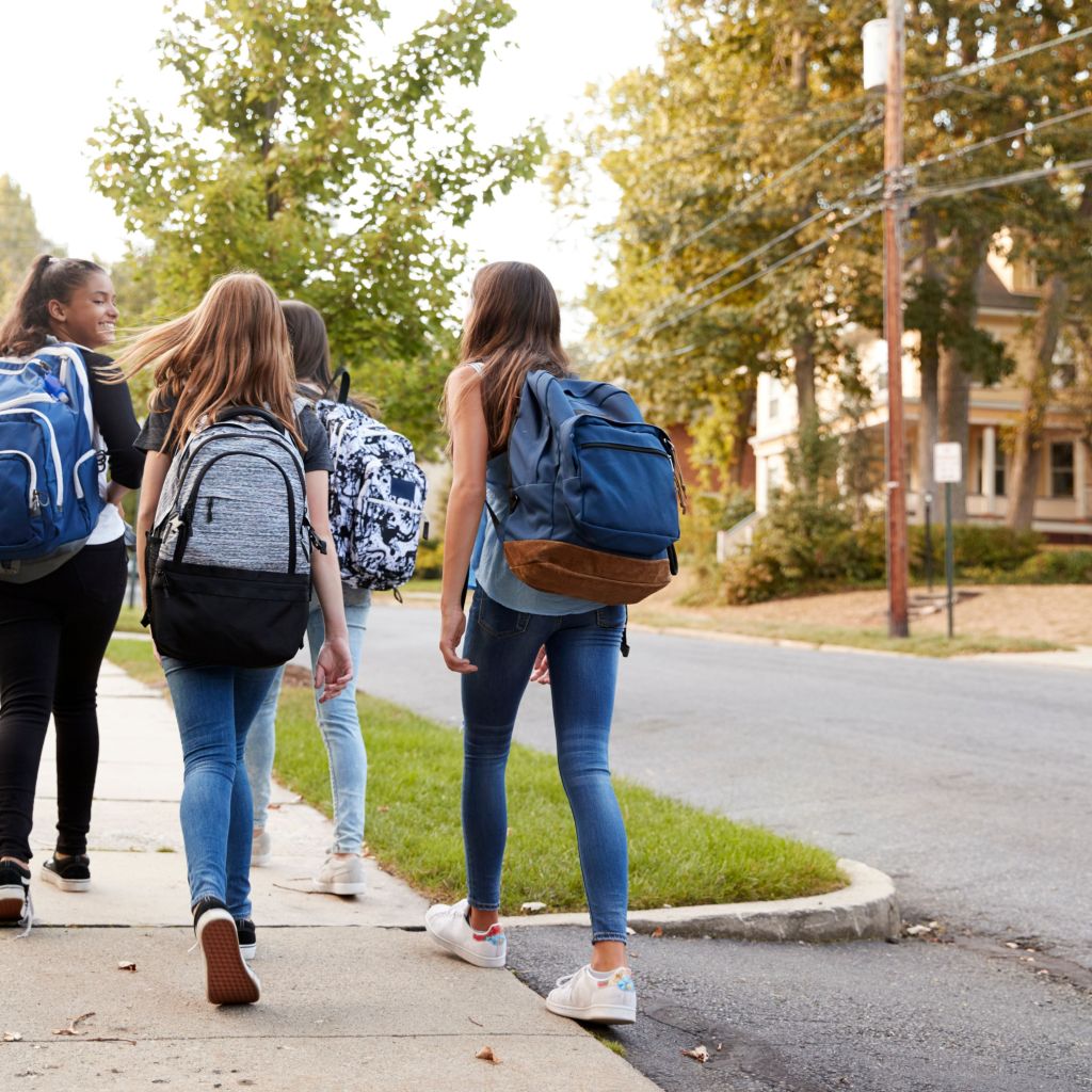 A group of teenage girls with backpacks walking down a suburban sidewalk, heading toward school or another destination, surrounded by trees and residential houses.