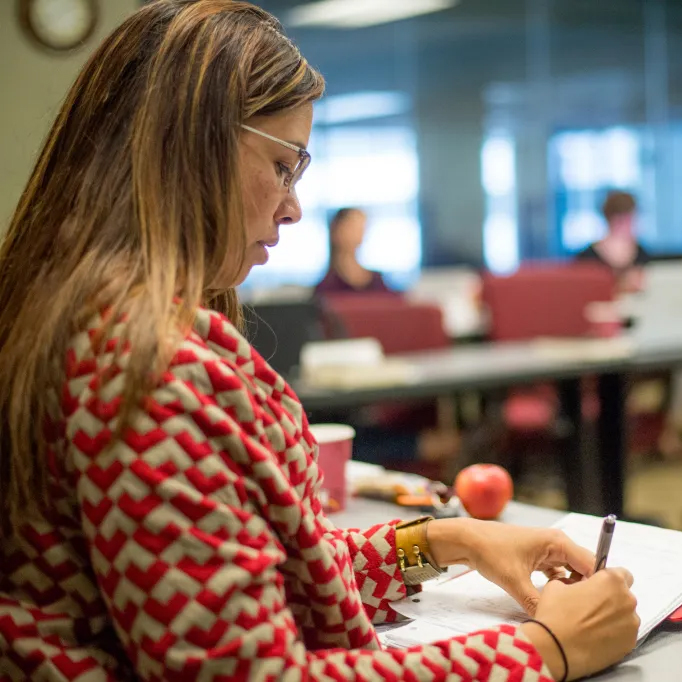 A woman is seated in a classroom or meeting room, attentively writing notes, with a cup and an apple on the table in front of her, suggesting she is engaged in a lecture or workshop setting.