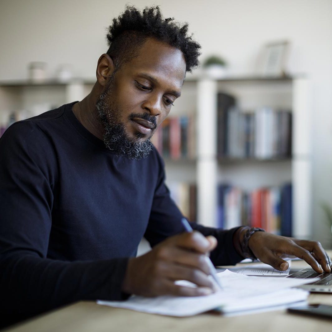 A man is sitting at a desk with a computer on his left and a stack of papers on his right with a pen in his right hand.