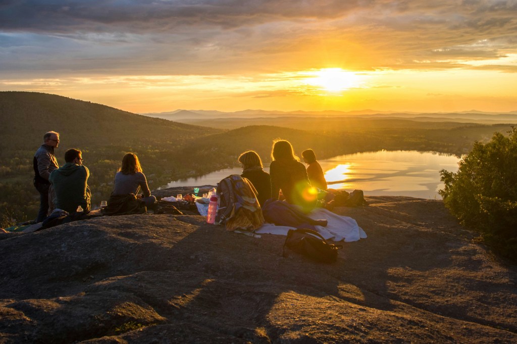 A group of people having a picnic at sunset with a view of a lake and mountains.